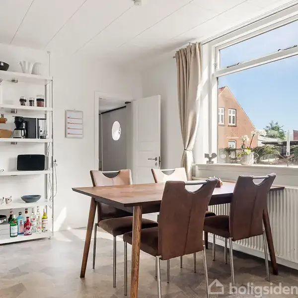Et spisebord med fire stole står i et lyst rum ved et vindue med gardiner, omgivet af en reol med køkkenredskaber. Ordet "BAR" ses på væggen.