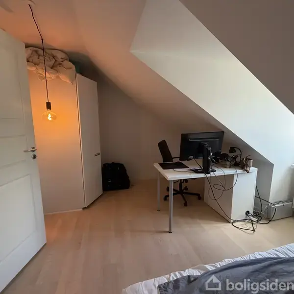 Et skrivebord med computer står under et skråtag i et værelse. Til venstre ses et klædeskab og en tændt loftslampe. Der er et tæppe på gulvet.