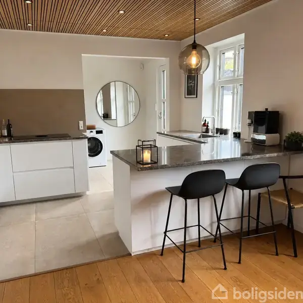 Køkken med moderne hvidt interiør og grå bordplade har to sorte barstole ved køkkenøen. En cirkulær vægspejl og vaskemaskine er synlige i baggrunden. Lys hænger fra træloftet.
