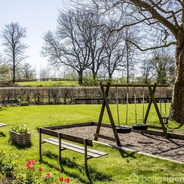 En legeplads med tre gynger står stille i en frodig have. Omgivet af træer, buske og blomsterbede, ses bænke langs kanten i solrigt vejr.