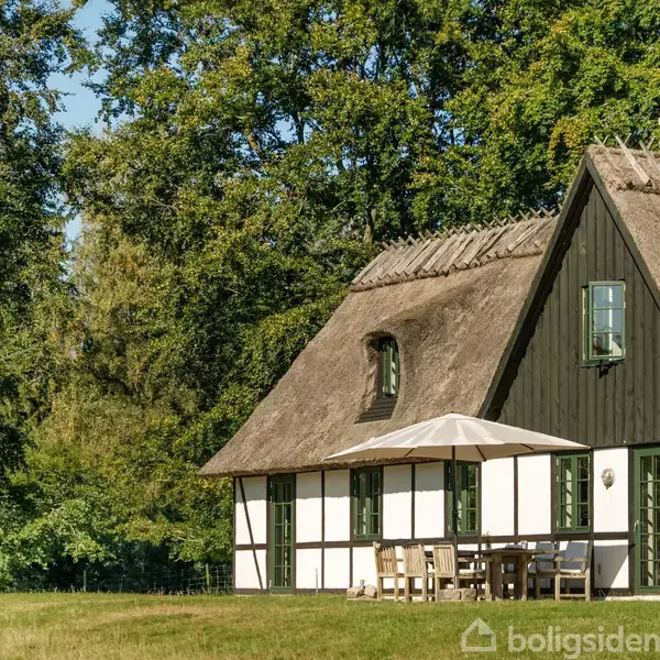 Stråtækt bindingsværkshus med sort-hvid facade står i solskinsvejr. En parasol og havemøbler ses udenfor. En flagstang med Dannebrog er placeret i haven omkranset af træer.