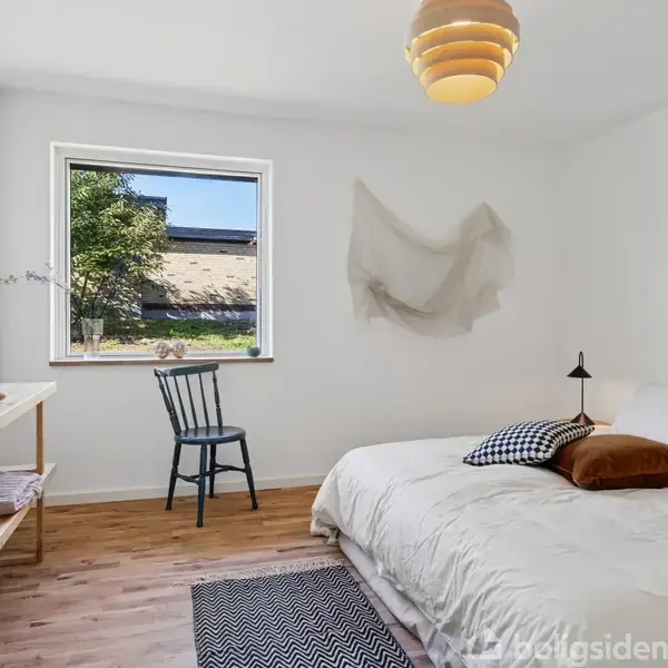 Soveværelse med en seng dækket af puder og en lampe over. En stol står ved et vindue med udsigt til det fri, og et træbord står ved siden af.