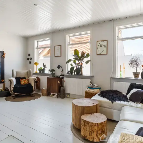 En lys stue med hvide vægge og lofter. En hvid sofa med puder står til højre, to træstubbe fungerer som borde. En lænestol med puder står ved vinduet.