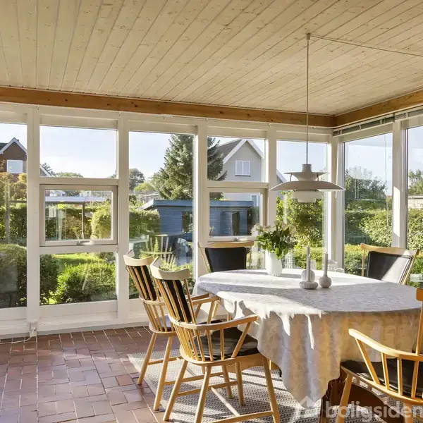 En spisestue med træstole omkring et dækket bord, omgivet af store vinduer, der giver udsigt til en have og nabohuse. Rummet har en træloft og murstensgulv.