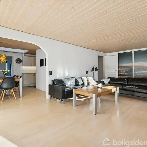 Stue med sorte lædersofaer og sofabord i forgrunden. I baggrunden en buegang, der fører til en spisestue med et bord og stole. Lyse trægulve og loft.