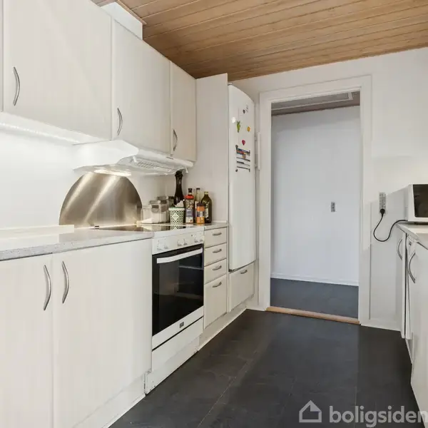 Et moderne køkken med hvide skabe. En ovn og køkkenredskaber står på bordpladen. Der er lyse fliser på gulvet. Døråbning fører til en tilstødende gang.