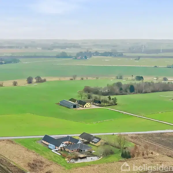 Luftfoto af en gårdbygning omgivet af åbne, grønne marker. Gården består af flere bygninger med en vej i forgrunden. Vindmøller ses i det fjerne på horisonten.
