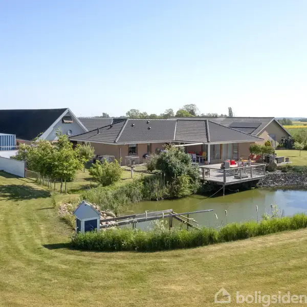 Hus med skråt tag står ved siden af en lille sø og træterrasse i en stor, grøn have. Landskabet er landligt med marker i baggrunden under en klar blå himmel.