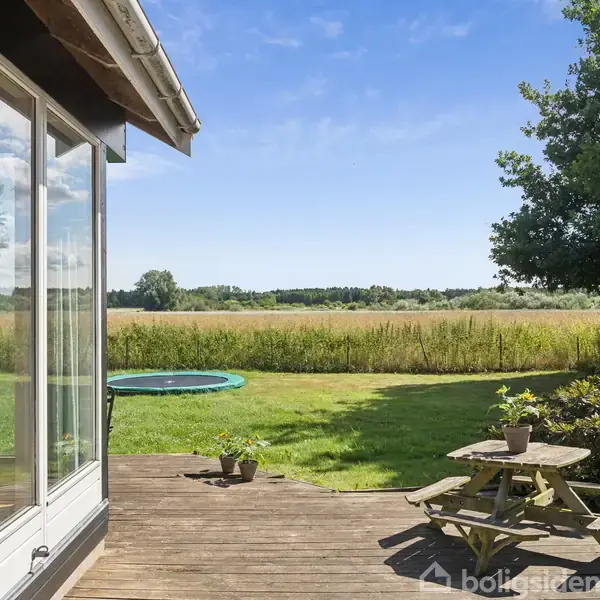 Et træhavebord med stole står på en terrasse ved siden af et hus. En stor græsplæne med en trampolin strækker sig ud mod et åbent marklandskab under en klar blå himmel.