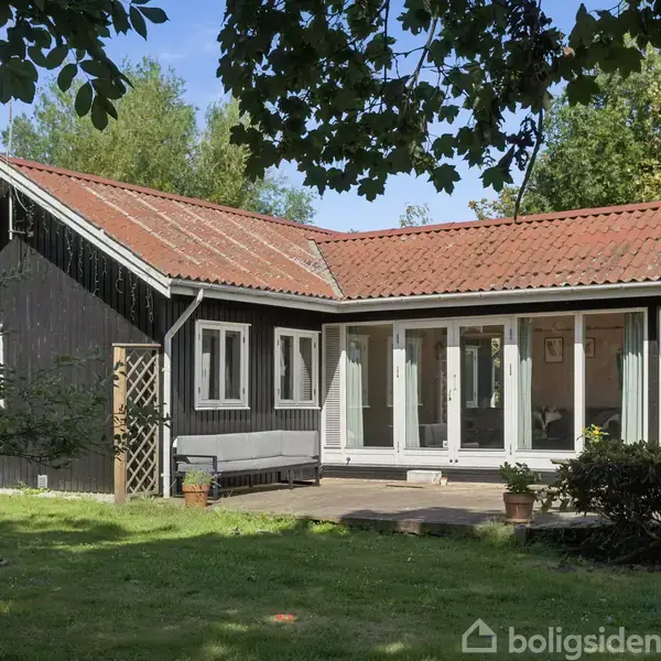 Et sortmalet sommerhus med hvidrammede vinduer og rødt tegltag står på en græsplæne omgivet af træer. På terrassen står et par blomsterkrukker.
