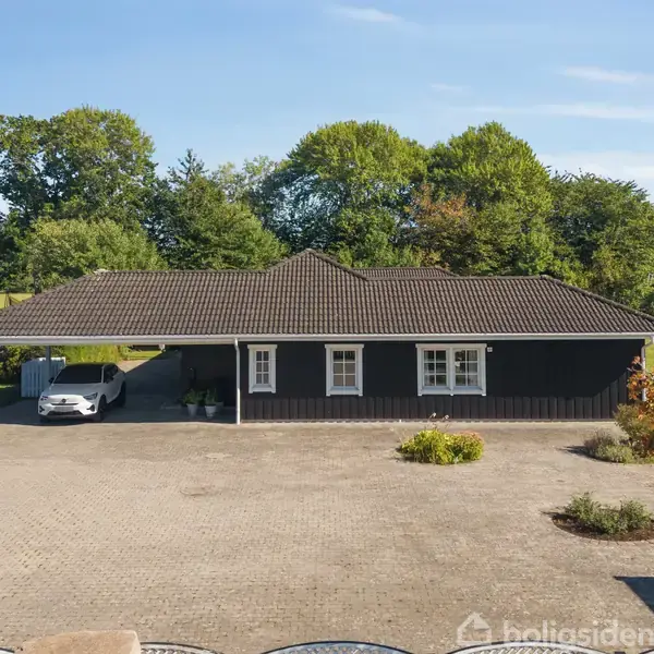 Et sort hus med saddeltag og flere vinduer står omgivet af tæt bevoksede grønne træer. En bil holder parkeret under en carport til venstre på en stor stenbelagt gårdsplads.
