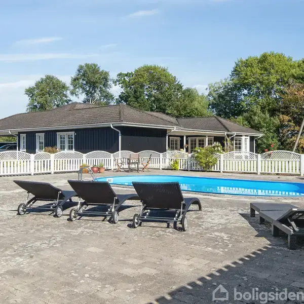 En swimmingpool omgivet af flisebelagt terrasse med fire liggestole står foran et mørkt træhus med hvidt hegn i en have med grønne træer i baggrunden.