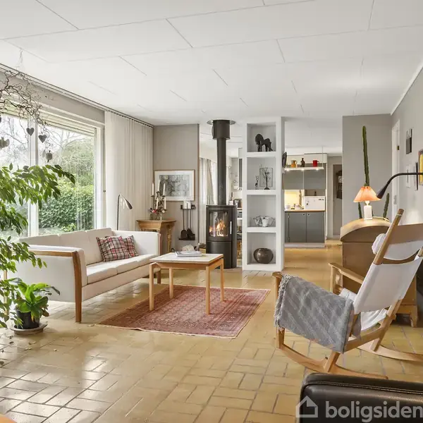 Stue med sofa og lænestol centreret om et sofabord, pejs brændende i baggrunden; store vinduer giver udsigt til have, moderne hyldesystem deler rummet, køkken ses i baggrunden.