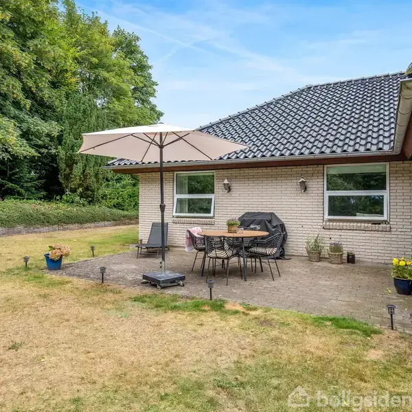 En terrasse med havebord, stole og en stor parasol står ved et murstenshus. Haven er omgivet af græsplæne og træer i baggrunden.