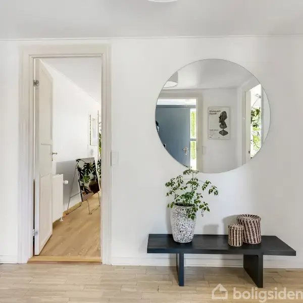 Rundt spejl hænger på en hvid væg, ovenover en sort bænk med to planter. Rummet er lyst med trægulve, og åbne døre fører til andre værelser.