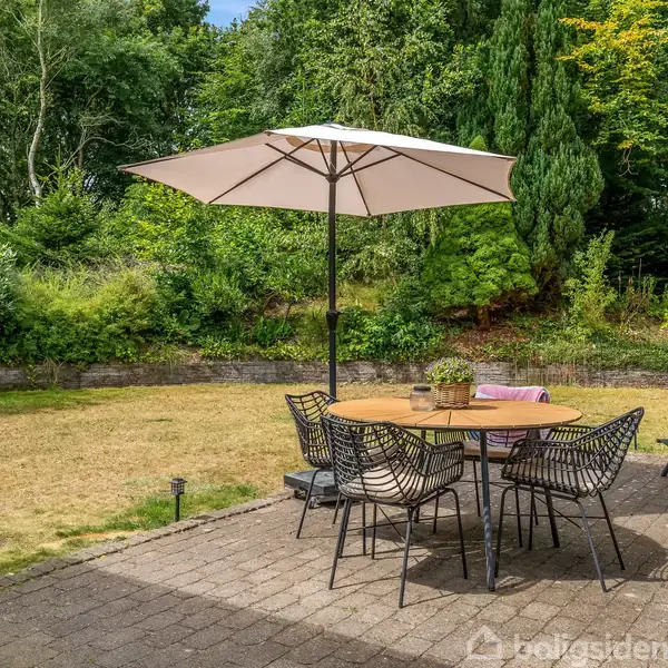 En træterrasse med et rundt bord og fire stole under en stor parasol. Omkring terrassen er en have med tæt beplantning og grønne træer.
