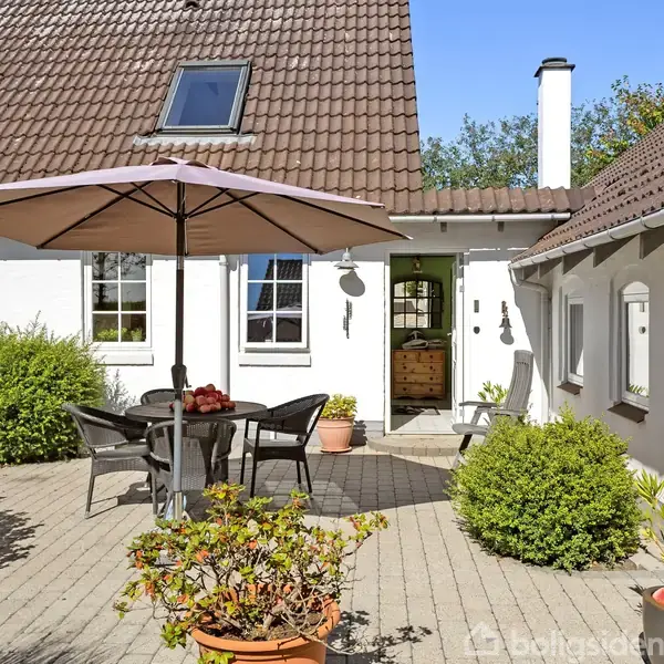 Udendørs gårdhave med spisebord og stole under en parasol. Hvide vægge og brune tegltage omgiver området. Grønne buske og planter i krukker skaber en hyggelig stemning.