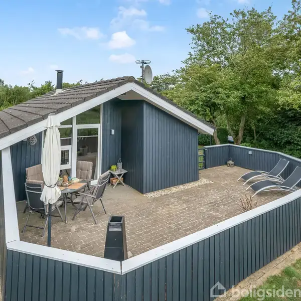Et træsommerhus i mørkeblå med en åben terrasse, hvor der står havemøbler og liggestole. Omgivet af græsplæne og træer under en blå himmel.