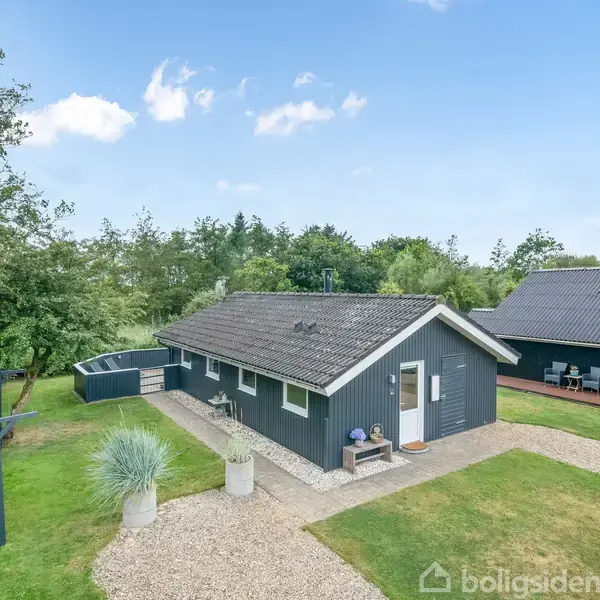 Et sortmalet hus med gråt tag står stille i en grøn have med græs og nogle træer. Huset har en lille terrasse og grussti foran.
