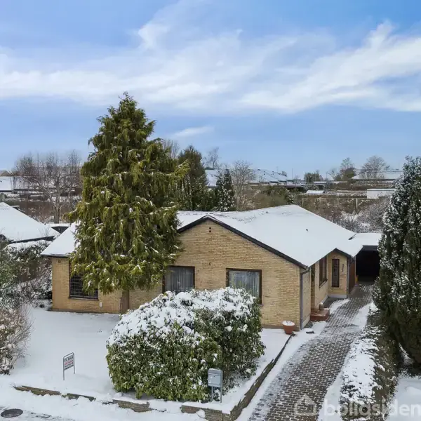 Et enkelt hus med fladt tag står dækket af sne. Indkørslen er omgivet af buske og træer, der også er sneklædte. Baggrunden viser et villakvarter under en blå himmel.