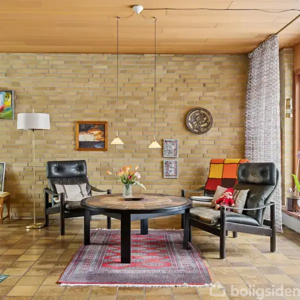 Stue med sorte lænestole står omkring et rundt træbord, dekoreret med blomster. Rødligt tæppe på gulvet. Murstensvægge med kunst, stor vindue med udsigt til snedækket have.