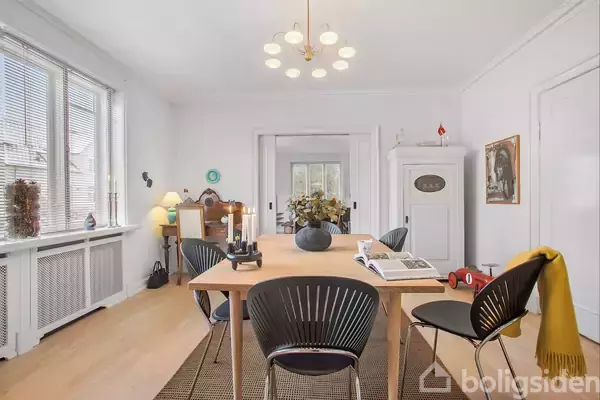 Et spisebord står i en lys spisestue med sorte stole omkring det. Bordet har bøger og en vase som dekoration, mens vinduerne ved siden slipper dagslys ind.