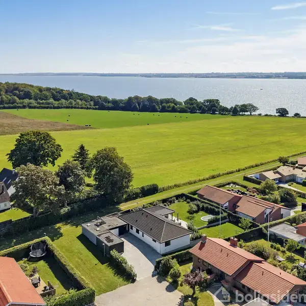 Boliger med røde tage ligger i række i et grønt boligområde. Bagved ses en stor græsmark tæt ved en skov, og i baggrunden er en stor sø eller fjord under en blå himmel.