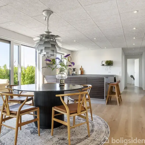 Rundt spisebord med fire træstole står på et tæppe i et lyst køkken-alrum. En moderne lampe hænger over bordet. Glasdøre i baggrunden åbner til grøn udsigt.