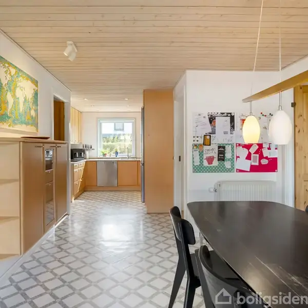 Et moderne køkken-alrum med et mørkt spisebord og sorte stole. Der hænger to hvide lamper over bordet. Væggene har dekorative opslagstavler. Gulvet er mønstret med lyse fliser.