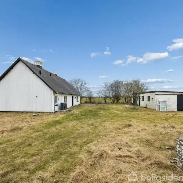Hus med hvidt træbeklædning og gråt tag står på en stor åben græsplæne. Ved siden af er en mindre bygning. Træer og marker udgør den landlige baggrund under en blå himmel.