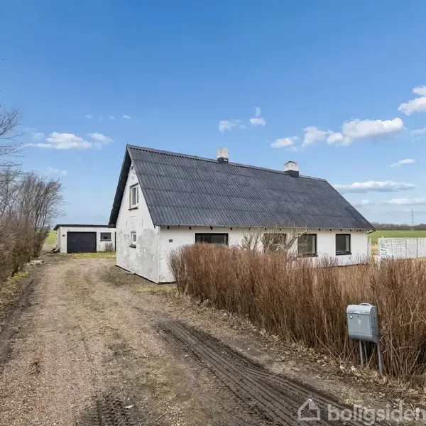 Hus med sort tag står stille ved grusvej, omringet af marker og træer i landligt område. En mindre bygning ses i baggrunden. Skyfri himmel ovenover.