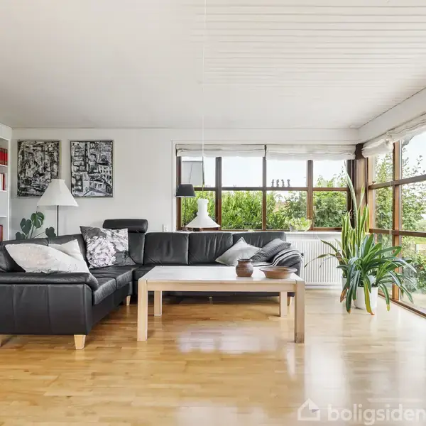 En sort lædersofa står i en lys stue ved siden af et trækaffebord. Bagved er en stor reol med bøger, lamper samt grønne planter ved vinduerne, der giver udsigt til terrasse.