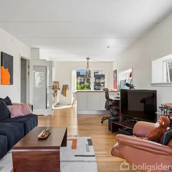 Stue med sort sofa og brun lænestol, hvor en brun bamse sidder. Et TV står på et bord. I baggrunden ses et køkken med hvide skabe og store vinduer, der giver naturligt lys.