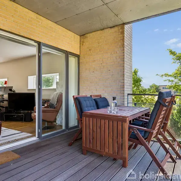 En udendørs terrasse med et træbord og fire stole, står i forbindelse med en lejlighed via glasdøre. Omkring er der en glasrækværk og grønne træer ses i baggrunden.