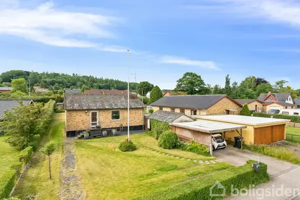En murstenshus med tilhørende garage og carport står på en grøn græsplæne omgivet af buske og træer. Omkringliggende huse og en skov kan ses i baggrunden under en blå himmel.
