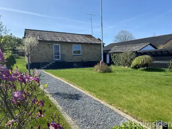 En enkeltstående murstensbygning står roligt, omringet af en velholdt græsplæne og blomstrende buske, med en grussti, der fører op til hoveddøren under en klar blå himmel.