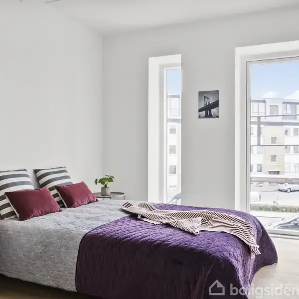 Soveværelse med dobbeltseng dækket af lilla sengetæppe og stribede puder. Ved siden af sengen står et lille bord med lampe, planten ved vindue, udsigt til bygning udenfor.