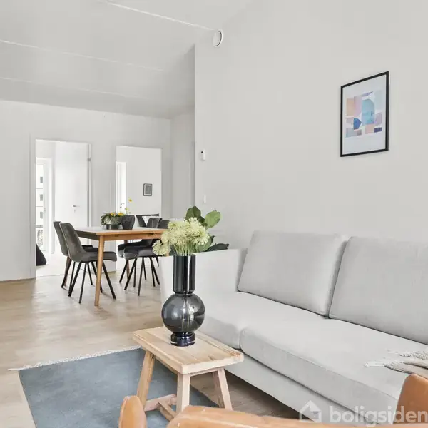 En lys stue med en grå sofa og en vase med blomster på et lille bord. I baggrunden står et spisebord med stole. Der hænger et billede på væggen.