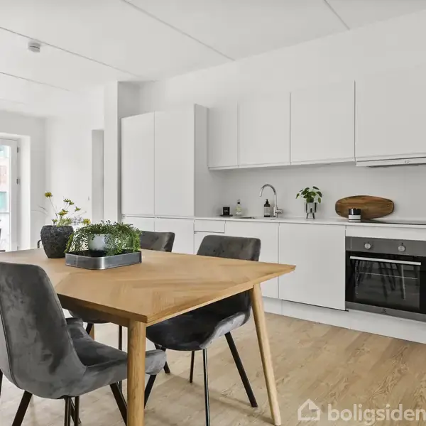 Spisebord med fire polstrede stole står midt i et moderne køkken. Rummet har hvide skabe, en indbygget ovn og er dekoreret med planter og en skærebræt på bordpladen.