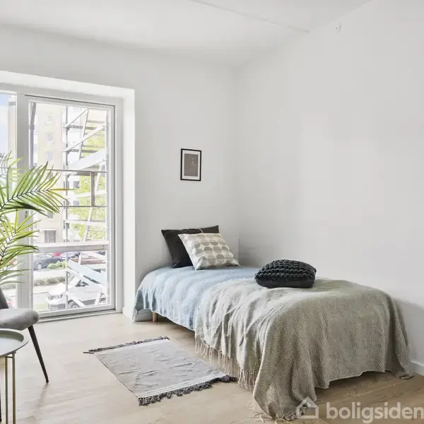 En enkeltseng står ved en væg med puder og et tæppe, ved siden af et stort vindue. En stol og et lille rundt bord med kopper er nærved.