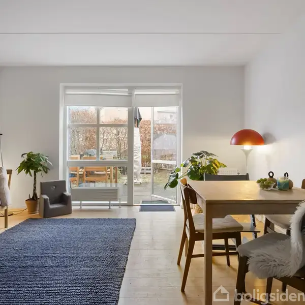 En stue med en sofa og lænestol til venstre, et spisebord med stole til højre, og store glasdøre i baggrunden, der åbner ud til en have. En blå løber dækker gulvet. Ingen tekst fundet.