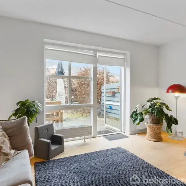 En lys stue indeholder en sofa med puder, en lænestol samt et skrivebord. Store vinduer åbner ud til en terrasse med udsigt til en have.