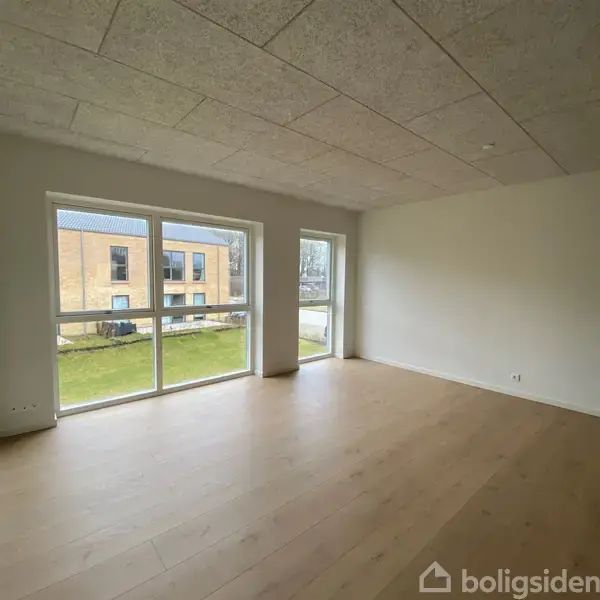 Et tomt værelse med trægulv og store vinduer, der lukker lys ind. Udenfor ses en græsplæne og en teglbygning i et boligområde.