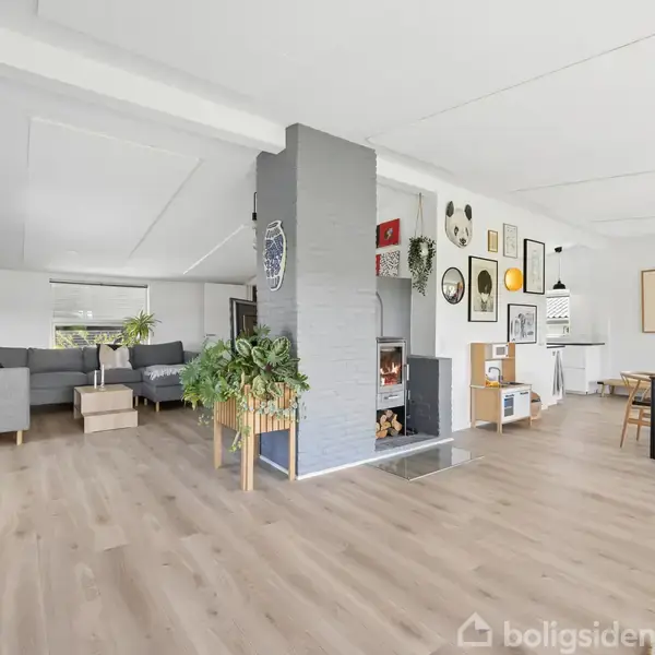 Stue med lys træparket, en moderne pejs i midten, grå sofaer til venstre, og et spisebord til højre. Væggen er dekoreret med billeder og kunstværker.