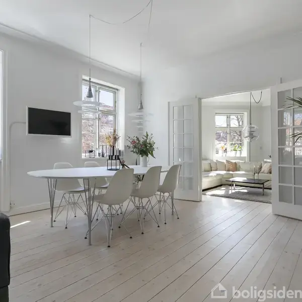 Spisebord med seks stole står i et lyst rum, omgivet af vinduer med udsigt til rød murstensbygning. I baggrunden ses stue med sofagruppe.
