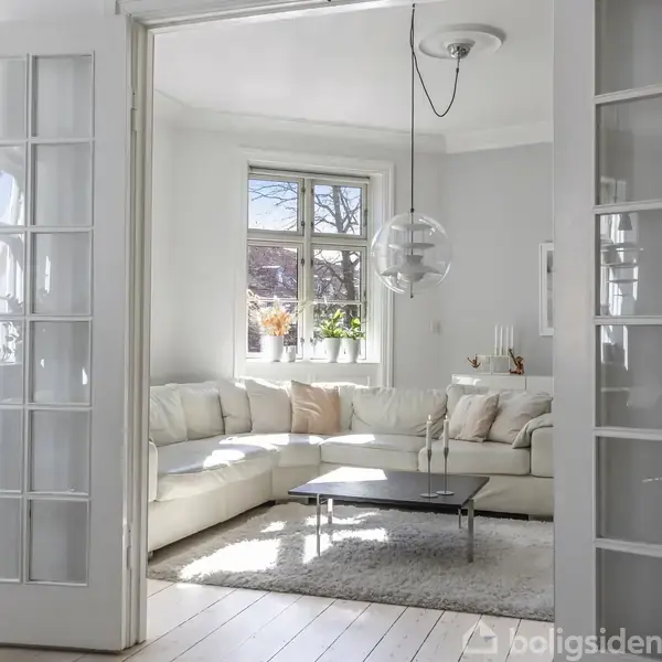 Hvid sofa står i en lys stue ved et vindue med planter. Ved sofaen er et sort sofabord og en lampe hænger fra loftet. Rummet er stilrent indrettet.