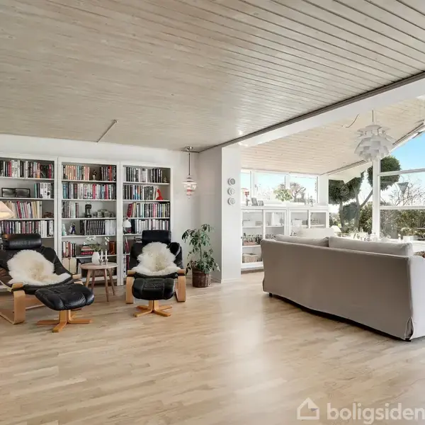 Et lyst, åbent stueområde med træloft og gulv. To lænestole står foran en reol fyldt med bøger. En stor sofa vender mod en terrasse med udsigt over haven.