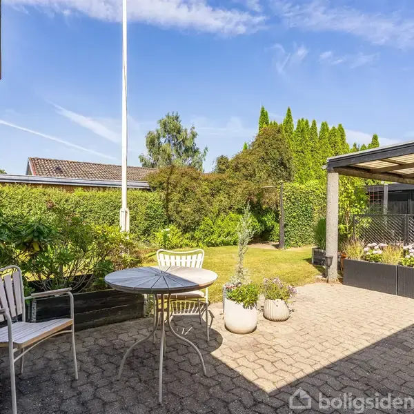 En terrasse med to hvide stole og et rundt bord står på en flisebelagt overflade. Baggrunden består af grønne buske og en pergola under en klar blå himmel.