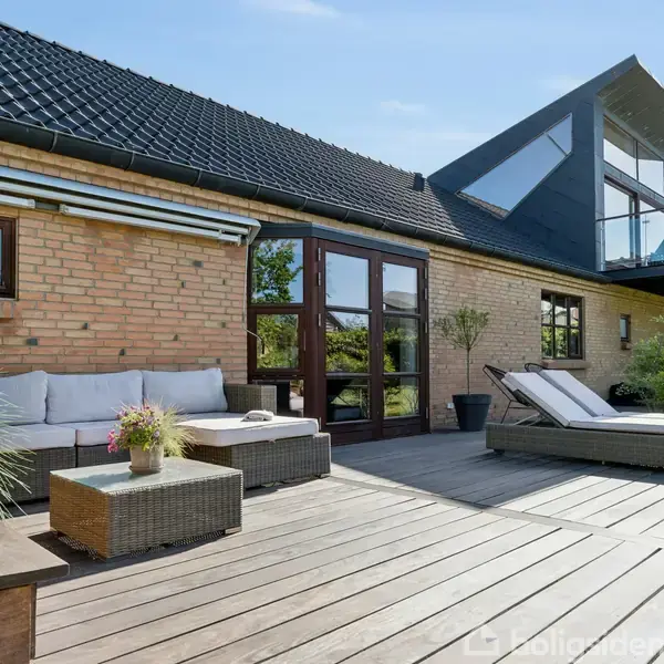 En moderne terrasse med sofagruppe og solseng står ved et murstenshus med store vinduer, bredt tag og havudsigt. Trædæk kantet af planter.