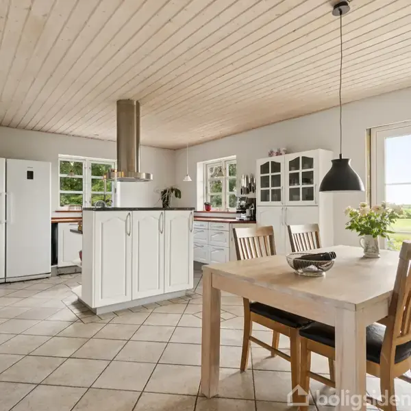 Et lyst køkken-alrum med træloft og flisegulv. Et stort spisebord er omringet af stole. Moderne elementer inkluderer et hvidt køkken med en emhætte. Store vinduer giver udsigt til naturen.